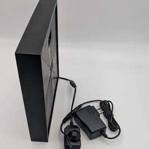 May include: A black square frame with a black and white image inside. The frame is connected to a black power adapter and a black dimmer switch with a knob.