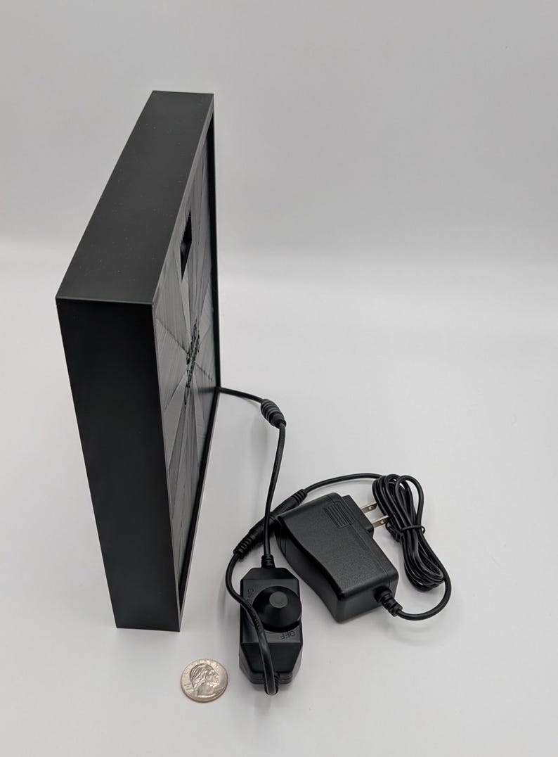 May include: A black square frame with a black and white image inside. The frame is connected to a black power adapter and a black dimmer switch with a knob.