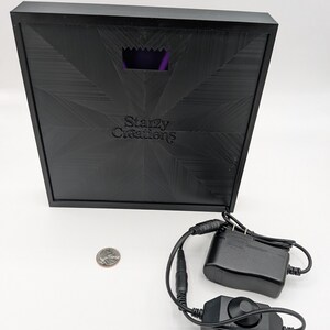 May include: A black square box with a purple light inside. The box has the text "Starzy Creations" on it. The box is connected to a black power adapter and a black dimmer switch.