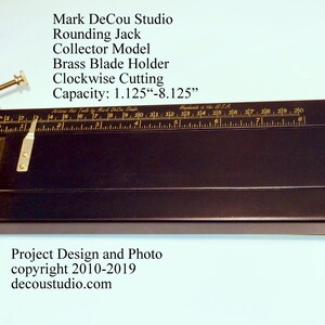 May include: A black wooden rounding jack with a brass blade holder and a ruler marked in inches. The jack is designed for clockwise cutting and has a capacity of 1.125 to 8.125 inches. The text "Mark DeCou Studio" is engraved on the jack.