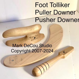 Op de afbeelding: Houten voetgereedschap voor het corrigeren van bunions. Het gereedschap is halfrond en heeft een handvat. Het gereedschap is gelabeld "Foot Tolliker Puller Downer Pusher Downer" en "Mark DeCou Studio Copyright 2007-2024".