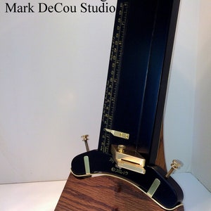 May include: A black and gold metal blade holder with a wooden base. The blade holder has a ruler on the side and a small brass plate with the text "Mark DeCou Studio" and "2019". The base is made of dark wood with a grain pattern. The text "Project Design and Photo copyright 2010-2019 decoustudio.com" is printed on the base.