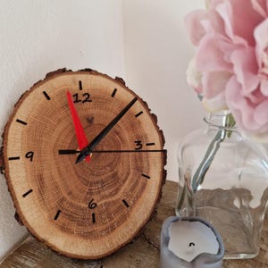 Könnte beinhalten: Eine runde Holzuhr mit natürlicher Maserung. Das Zifferblatt hat schwarze Ziffern und Zeiger, mit einem roten Sekundenzeiger. Die Uhr steht auf einem Holzregal, neben einer Kerze und einer Vase mit rosa Blumen.