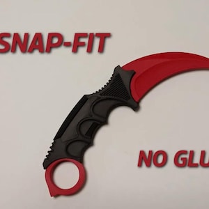 Puede incluir: Un cuchillo karambit con una hoja roja curvada y un mango negro. El mango tiene un anillo para el dedo y cortes decorativos. Las palabras "SNAP-FIT" y "NO GLUE" están en texto rojo.