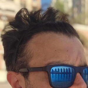 May include: Black sunglasses with blue mirrored lenses reflecting a waterfront scene. The sunglasses have a dark frame and are worn by a person with dark hair. The image is a close-up of the face.