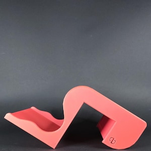 Può includere: Un supporto per telefono in plastica rosso vivo con un design curvo. Il supporto ha un piccolo ritaglio rettangolare sul lato.