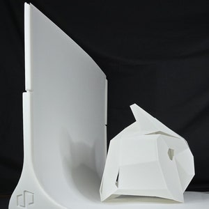 May include: A white, curved, backdrop with a white, geometric, abstract sculpture in front of it. The backdrop has a logo on the bottom left corner.