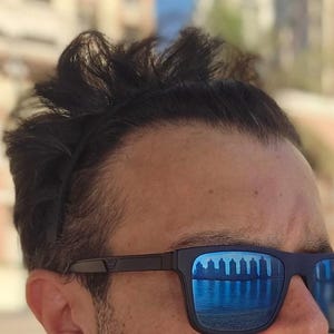 May include: Black sunglasses with blue mirrored lenses reflect a waterfront scene. The sunglasses have a dark frame and are worn by a person with dark hair. The image is a close-up of the person's face.