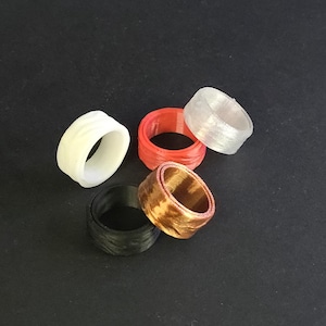 May include: A collection of five rings in various colors and textures. The rings include white, red, clear, black, and bronze. The rings are made of a 3D printed material and are displayed against a dark background.