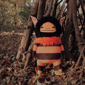 May include: A plush toy with black fur, orange fur trim, and black and orange stripes. The toy has large, black eyes and a friendly smile. The toy is standing in a woodland setting.