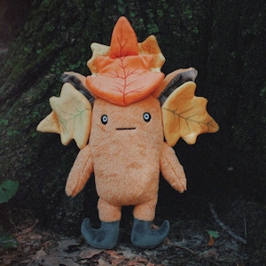 May include: A plush toy with a brown, furry body and large, orange leaf ears. The toy is wearing a hat made of a single orange leaf and has black shoes.