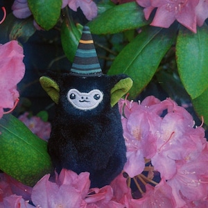 May include: A black plush toy with a white embroidered face and green ears, wearing a striped hat. The toy is surrounded by pink rhododendron flowers and green leaves, creating a whimsical scene.