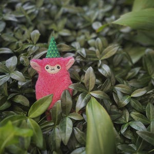 May include: A plush pink creature with large ears and a small face, wearing a green cone-shaped hat with white polka dots. The stuffed animal is surrounded by green leaves, creating a natural, whimsical scene.