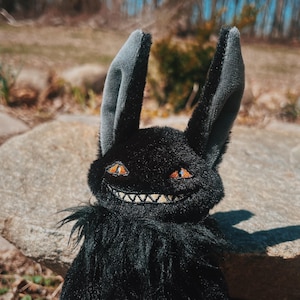 Puede incluir: Un conejo de peluche negro con largas orejas forradas en gris y ojos naranjas. El conejo tiene una amplia sonrisa con dientes y un cuerpo negro y esponjoso. El peluche está sobre un fondo natural al aire libre.
