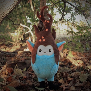 Puede incluir: Un juguete de peluche azul y marrón con una cabeza con forma de árbol y ojos grandes. El juguete está de pie sobre un lecho de hojas marrones.