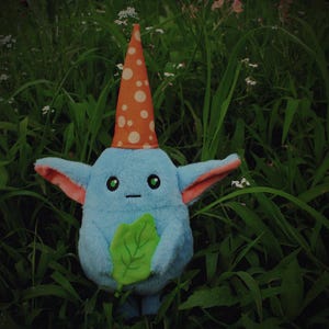 May include: A plush blue creature with a pointed orange hat with white polka dots, holding a green leaf. The creature has pink inner ears and green eyes. It is set against a background of green grass and small white flowers.