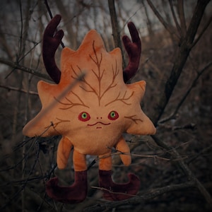 以下が含まれることがあります： 顔と角のある葉の形をしたぬいぐるみ。おもちゃはオレンジと茶色で、緑色の目と笑顔の口をしています。おもちゃは枝の山の上に座っています。