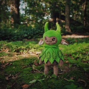 Könnte beinhalten: Eine Plüschpuppe einer Waldkreatur mit einem grünen, blattförmigen Outfit und einer passenden Kapuze mit Hörnern. Die Puppe hat ein braunes Gesicht mit gestickten Details und steht in einer moosigen Waldumgebung.