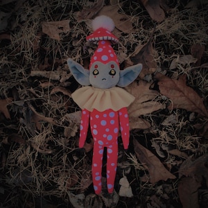 May include: A handmade plush toy elf with a red and white polka dot body, a yellow ruffle collar, and a mushroom hat. The elf has gray skin, large blue eyes, and a sad expression. The elf is lying on a bed of brown leaves.