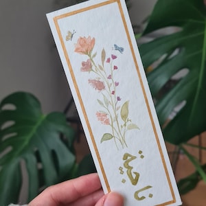 May include: A watercolour bookmark with a gold border featuring a floral design with pink, peach, and green flowers and leaves. Two butterflies are also painted on the bookmark. The text "يا قُتَيْلَ" is written in gold calligraphy at the bottom of the bookmark.