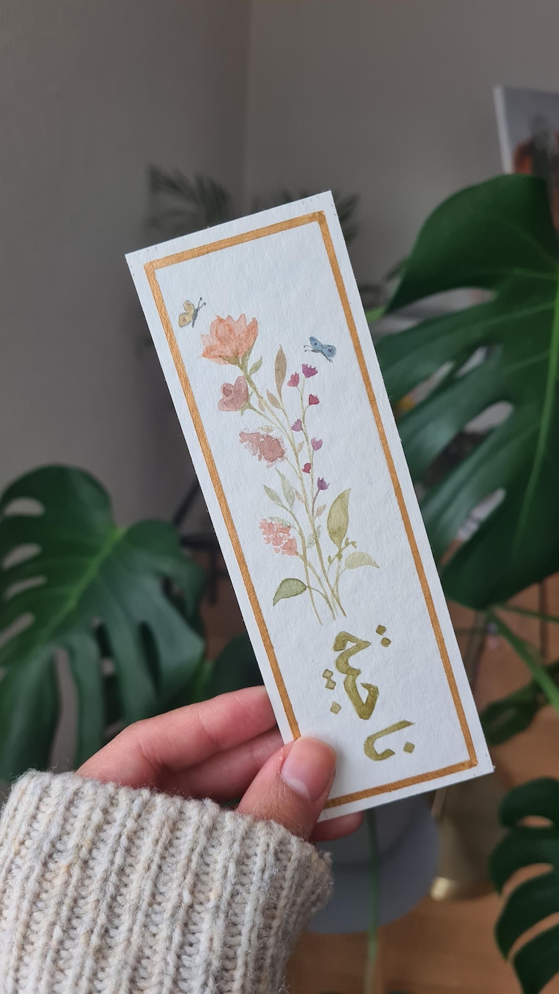 May include: A watercolour bookmark with a gold border featuring a floral design with pink, peach, and green flowers and leaves. Two butterflies are also painted on the bookmark. The text "يا قُتَيْلَ" is written in gold calligraphy at the bottom of the bookmark.