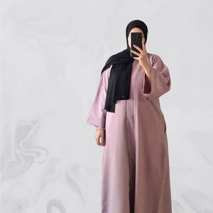 May include: A long-sleeved, dusty rose abaya with a black headscarf. The abaya has a long, flowing silhouette and a simple, elegant design.