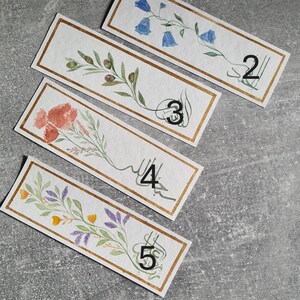 May include: Five watercolour bookmarks with gold trim and floral designs. Each bookmark has a number from 2 to 5 written on it.