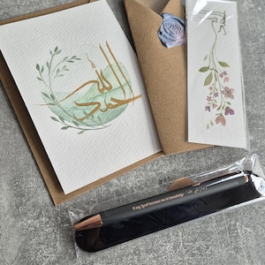 May include: A set of Islamic calligraphy greeting cards featuring a green and gold design. The cards are packaged with a brown envelope and a bookmark with a floral design. A black pen with gold lettering is also included. The pen has the inscription "O my Lord increase me in knowledge."