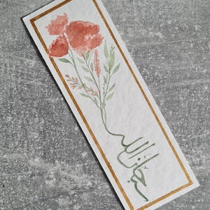 May include: Watercolor bookmark with a floral design. It features coral flowers, green leaves, and a gold border. The bookmark includes Arabic calligraphy in green ink. The background is a grey surface.