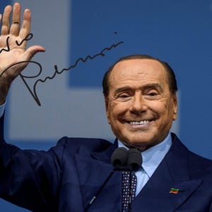 Puede incluir: Un hombre con un traje azul marino con una camisa azul claro y una corbata estampada sonríe mientras saluda. Tiene la mano levantada y hay una firma en su mano. El fondo es un degradado de azul.
