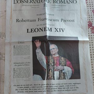 Può includere: Un giornale intitolato "L'OSSERVATORE ROMANO" con il titolo "HABEMUS PAPAM" e il nome "Robertum Franciscum Prevost". L'immagine presenta un uomo in abito religioso, che saluta. È presente anche del testo in italiano.