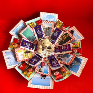 Może przedstawiać: Kolorowe pudełko eksplodujące z pluszowym misiem, tabliczkami czekolady i zdjęciami. Pudełko jest ozdobione czerwoną i białą kratą i ma wiele przegródek.