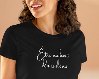 Op de afbeelding: Zwart T-shirt met de Franse zin "Être au bout du rouleau" in witte letters. Het shirt heeft een ronde hals en wordt gecombineerd met een blauwe spijkerbroek. Het model lacht.