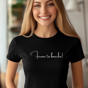 Puede incluir: Camiseta negra con la frase "Ferme ta bouche!" en escritura blanca. La camiseta es de estilo cuello redondo y parece estar hecha de un material suave. La modelo sonríe.