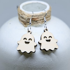 Op de afbeelding: Een paar bungelende oorbellen met lichtgekleurde houten spookbedels. Elk spookje heeft een lasergesneden ontwerp met een lachend gezicht, inclusief twee gebogen ogen en een brede mond. De bedels zijn bevestigd aan zilverkleurige oorhaken.
