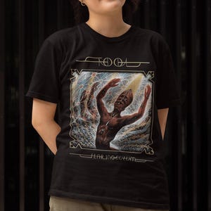 May include: Black t-shirt with a graphic design featuring a figure with outstretched arms, with the word "TOOL" at the top and "FEAR INOCULUM" at the bottom. The person is wearing tan cargo pants.