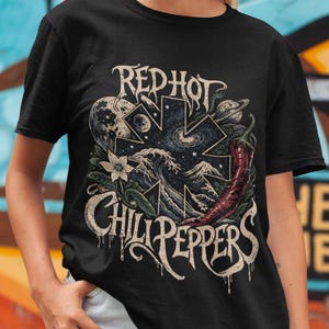 May include: Black t-shirt featuring the words "Red Hot Chili Peppers" in a stylized font, along with a graphic of a chili pepper, waves, and celestial elements. The shirt is worn by a person.