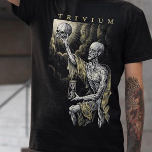May include: Black t-shirt with a graphic print of a skeletal figure holding an hourglass and a skull. The text "TRIVIUM" is printed in gold lettering on the shirt.