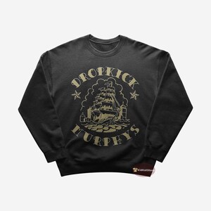 Peut inclure: Sweat-shirt noir avec un motif doré d'un navire et le texte "Dropkick Murphys" sur le devant.