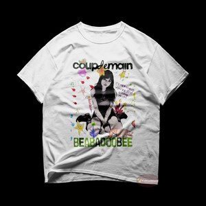 May include: White t-shirt featuring a black and white illustration of a woman sitting, with a colourful background. The text "coupdemain" and "BEABADOOBEE" are printed on the shirt.