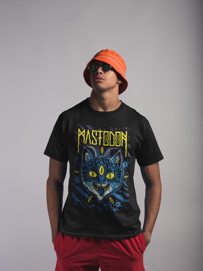 May include: A black t-shirt featuring a blue and yellow illustration of a cat with three eyes and the word "MASTODON" in yellow lettering.