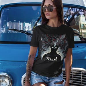Puede incluir: Una camiseta negra con un estampado gr&aacute;fico de una persona con un dise&ntilde;o en espiral sobre su cabeza. El texto "tool" est&aacute; impreso debajo del gr&aacute;fico. La persona lleva gafas de sol y pantalones cortos de mezclilla.