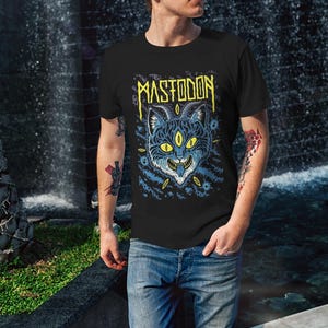 May include: A black t-shirt featuring a graphic print of a blue cat with yellow eyes and horns. The text "MASTODON" is printed in yellow above the cat.