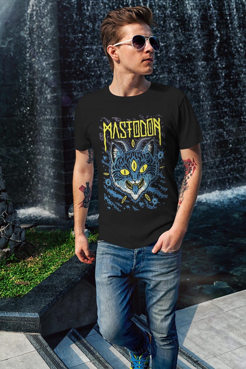 May include: A black t-shirt featuring a graphic print of a blue cat with yellow eyes and horns. The text "MASTODON" is printed in yellow above the cat.