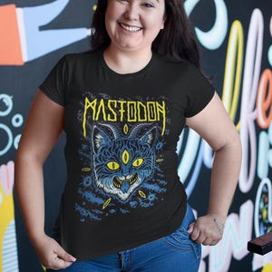 May include: A black t-shirt featuring a graphic of a blue cat with yellow eyes and the word "MASTODON" in yellow lettering.