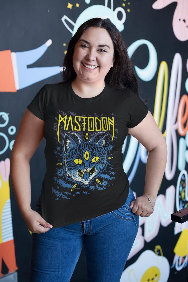 May include: A black t-shirt featuring a graphic of a blue cat with yellow eyes and the word "MASTODON" in yellow lettering.