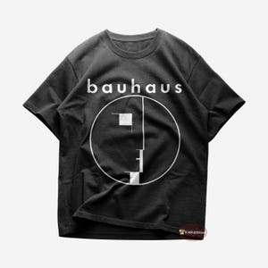 Puede incluir: Camiseta negra con un gráfico blanco de un círculo que contiene una cara estilizada y la palabra "bauhaus" encima.