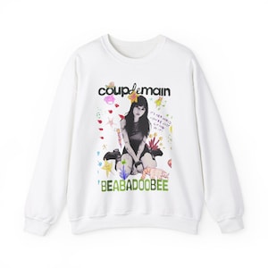 May include: White long-sleeved sweatshirt with a colourful graphic print featuring a girl sitting down with a cat. The graphic also includes the text "coupdemain" and "BEABADOOBEE".
