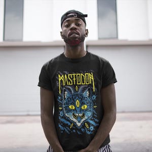 May include: A black t-shirt featuring a graphic print of a blue and yellow cat with three eyes and the word "MASTODON" in yellow lettering.
