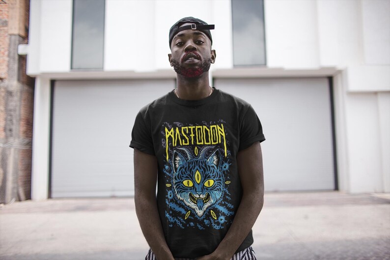May include: A black t-shirt featuring a graphic print of a blue and yellow cat with three eyes and the word "MASTODON" in yellow lettering.